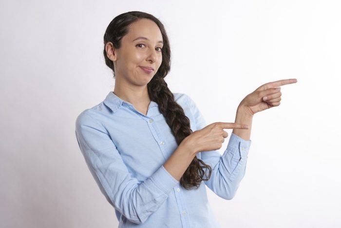 Une femme avec une longue tresse de cheveux noirs qui sourit en montrant la direction vers la droite avec ses deux mains