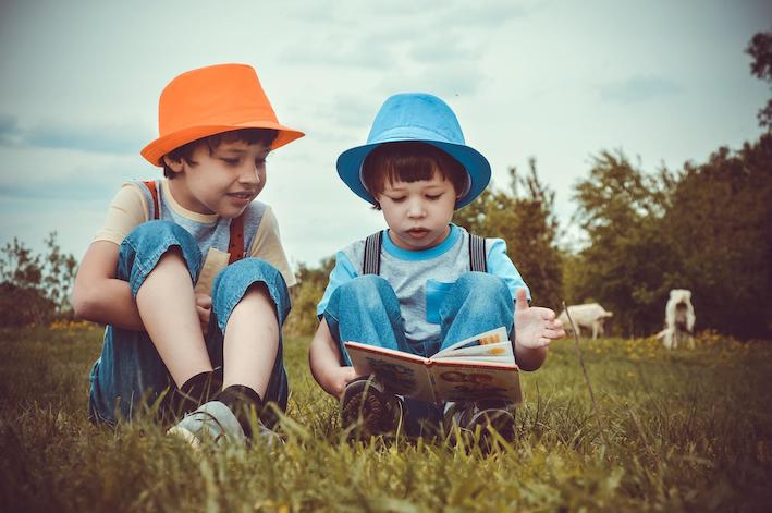 L'écoute active : deux petits garçons assis dans l'herbe l'un écoute attentivement avec un chapeau orange l'autre qui lit un livre lillustré et qui porte un chapeau bleu